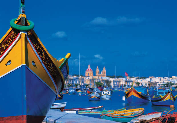 Colorful wooden boats float in a vibrant blue harbor, with a clear sky above. In the background, a church with twin towers stands amidst a town, creating a picturesque coastal scene.